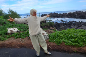 A person with short light-colored hair stands with arms outstretched on a coastal path overlooking a rocky shoreline and ocean under a cloudy sky.
