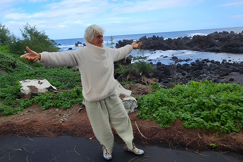 A person with short light-colored hair stands with arms outstretched on a coastal path overlooking a rocky shoreline and ocean under a cloudy sky.