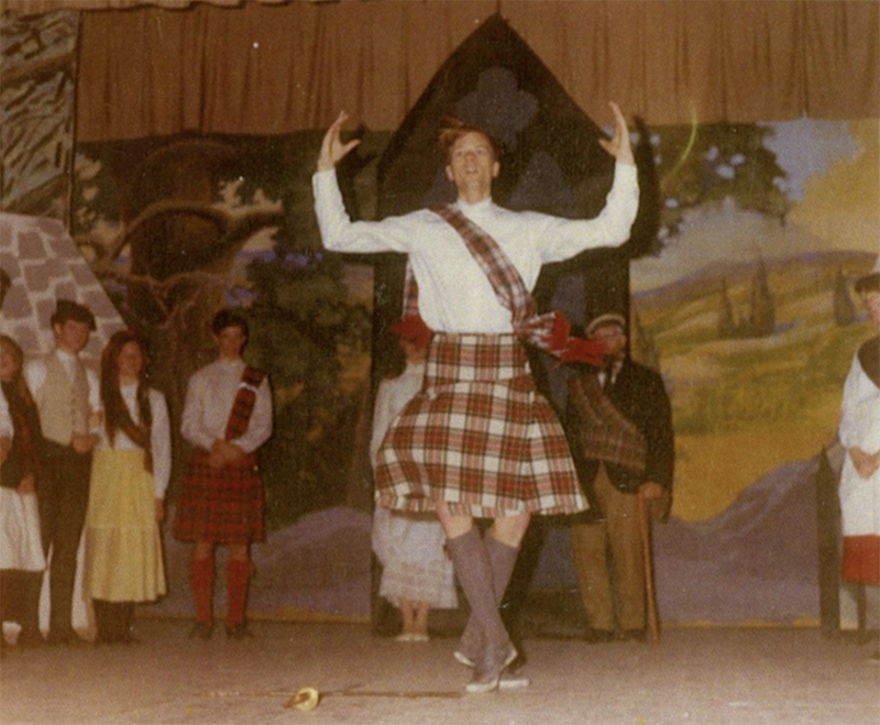 A performer in a plaid kilt-style costume dances center stage with raised arms during a theatrical production, while other cast members stand against a painted village backdrop.