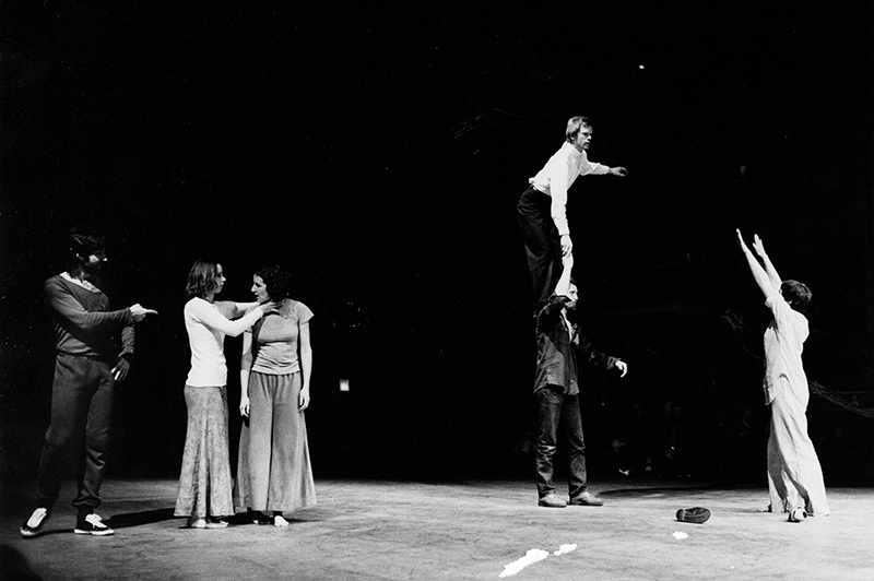 A black-and-white stage performance shows a group of dancers spread across a dark theater space, including one performer balancing atop another’s raised hand while others gesture and interact around them.