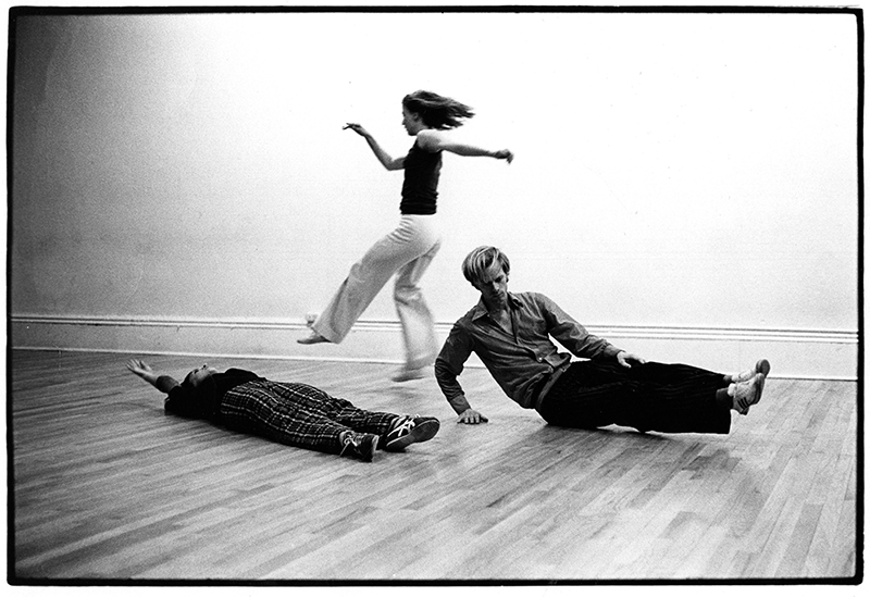 A black-and-white dance studio scene shows three performers interacting through contrasting movements, with one lying on the floor, one balancing in a seated pose, and one moving dynamically across the wooden floor.
