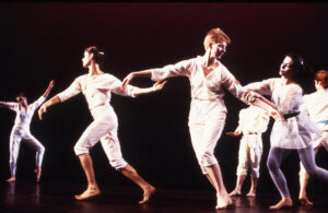 A group of dancers dressed in light-colored costumes perform interconnected movements across a dark stage, with several performers extending their arms and holding hands under warm theatrical lighting.