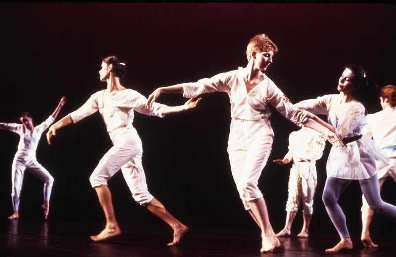 A group of dancers dressed in light-colored costumes perform interconnected movements across a dark stage, with several performers extending their arms and holding hands under warm theatrical lighting.