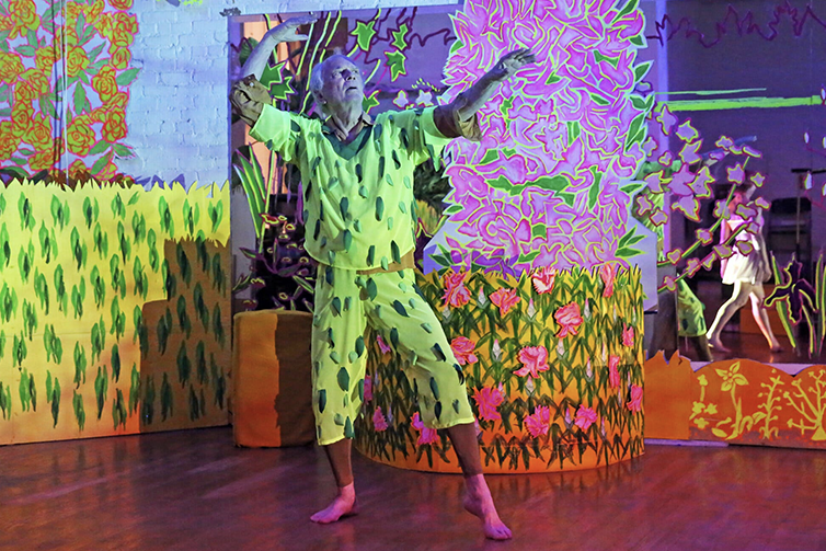 A barefoot performer in a bright green patterned costume poses with raised arms amid a vivid, colorful garden-themed installation filled with painted flowers, foliage, and neon-like decorative panels.