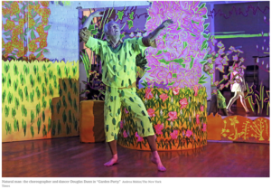 A barefoot performer in a bright green patterned costume poses with raised arms amid a vivid, colorful garden-themed installation filled with painted flowers, foliage, and neon-like decorative panels.