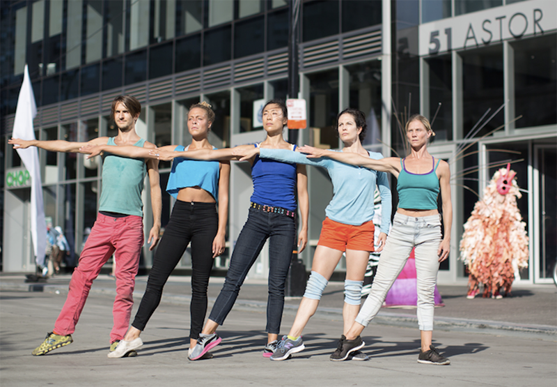 Five performers in casual, brightly colored clothing stand in a synchronized pose with outstretched arms in an urban plaza, with a modern office building and a costumed figure visible in the background.