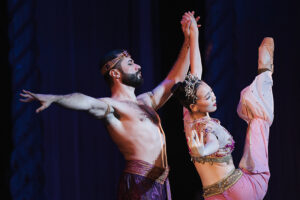 Ahmad and his partner in Arabian Nutcracker costumes execute a synchronized pose under theatrical lighting, with the partner extending a leg high in the air while both hold hands center stage.