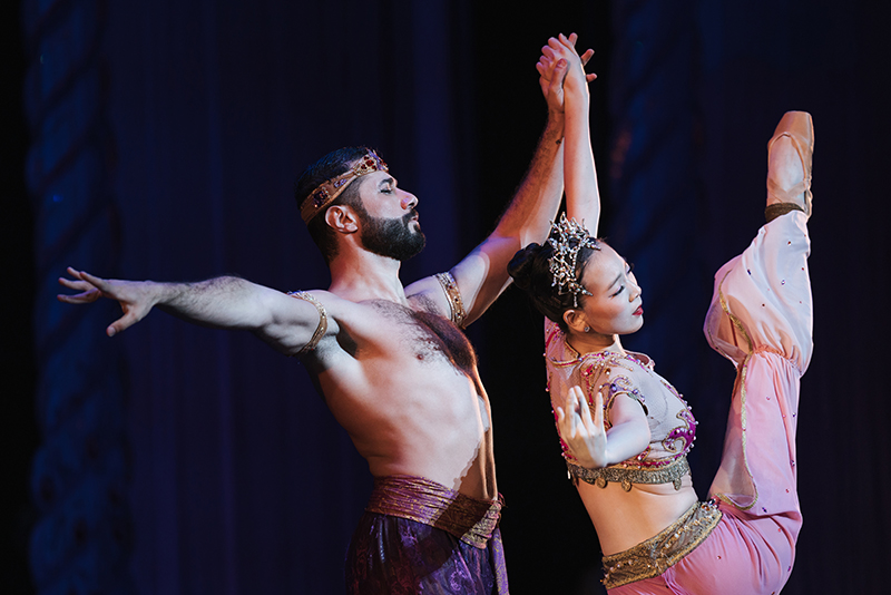 Ahmad and his partner in Arabian Nutcracker costumes execute a synchronized pose under theatrical lighting, with the partner extending a leg high in the air while both hold hands center stage.