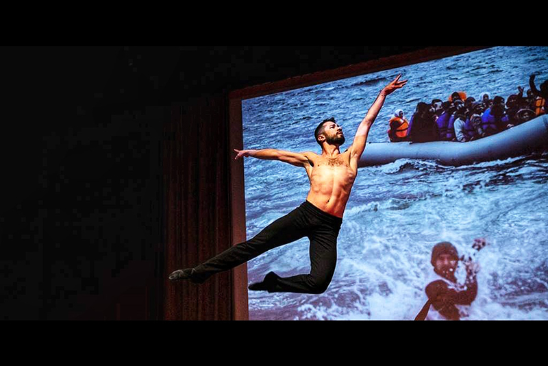 Ahmad performs a dramatic midair leap on a stage in front of a large projected image of people in an inflatable boat crossing rough ocean waters.