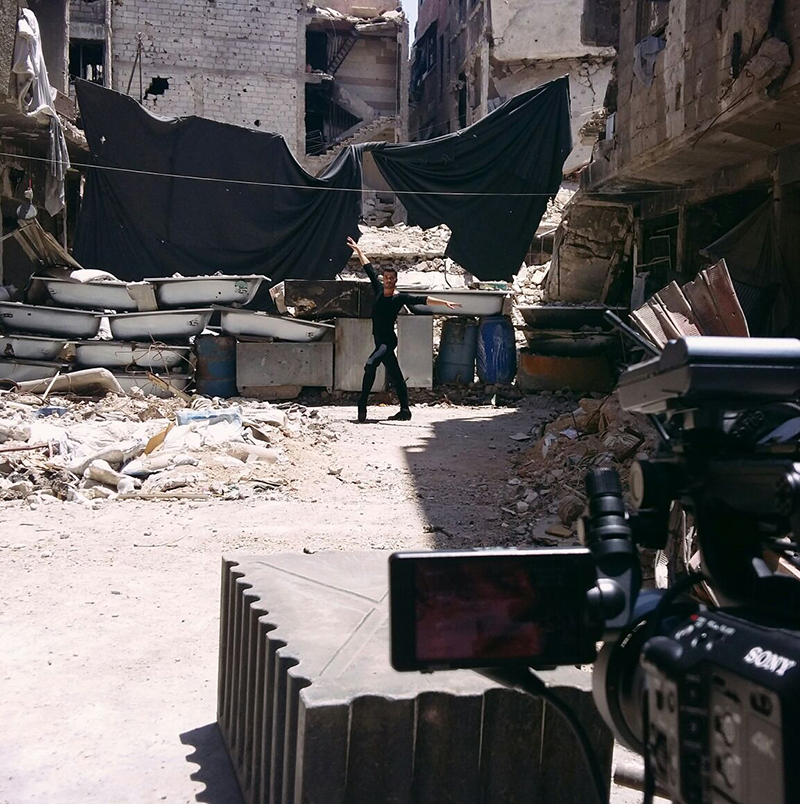 Ahmad poses in the middle of a rubble-strewn street between damaged buildings while a video camera films the scene in the foreground.