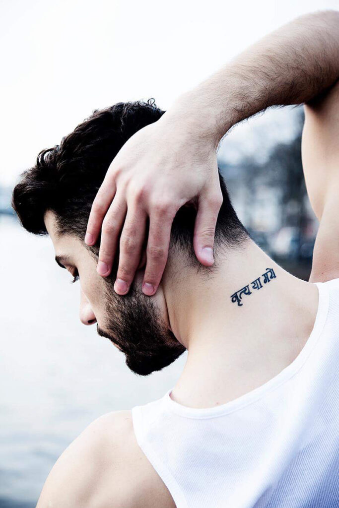 A close-up portrait oh Ahmad in a white sleeveless shirt touching the back of his neck, revealing a small tattoo in Devanagari script just below the hairline.