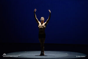 Ahmad stands alone under a spotlight on a dark stage, raising both arms in a poised ballet stance against a deep blue backdrop.