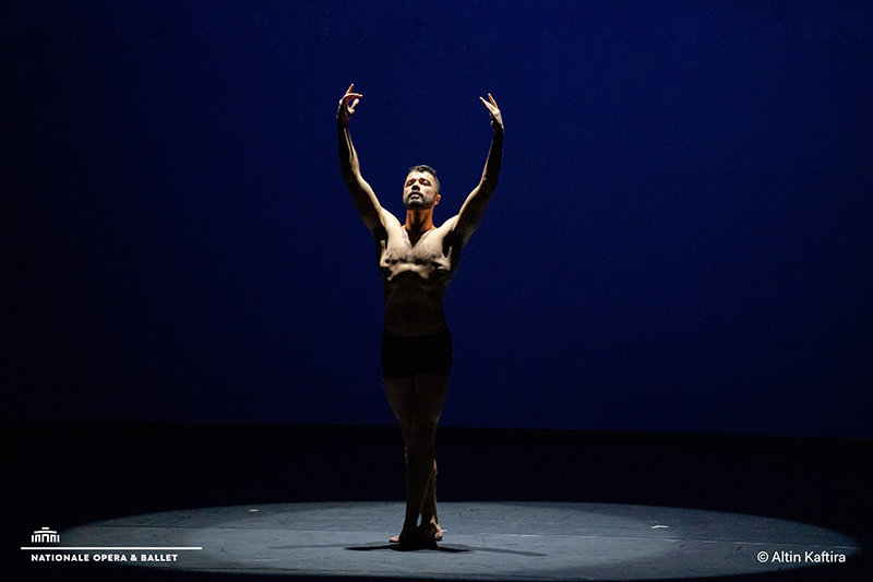 Ahmad stands alone under a spotlight on a dark stage, raising both arms in a poised ballet stance against a deep blue backdrop.