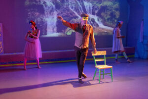 A theatrical performance unfolds on a softly lit stage, where three costumed performers stand before a projected waterfall backdrop, with a masked central figure posing beside a brightly painted chair under purple and blue lighting.