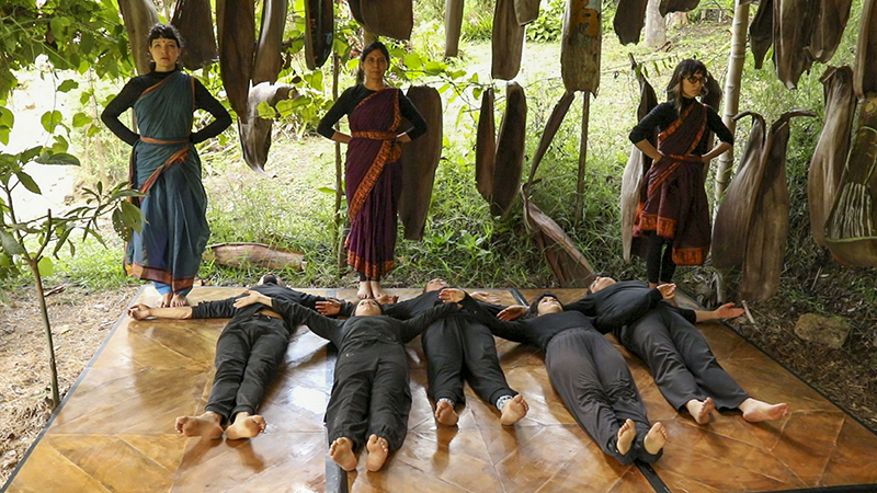 Three standing participants in colorful draped garments hold formal poses around a group of participants lying on a wooden platform beneath hanging natural plant structures in an open-air garden setting.