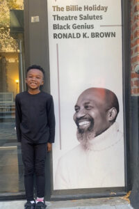 Copilot said: A child dressed in black stands beside a large theater display featuring a portrait of Ronald and the text, “The Billie Holiday Theatre Salutes Black Genius RONALD K. BROWN,” outside a building entrance.