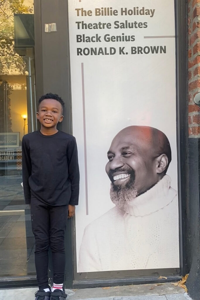 Copilot said:A child dressed in black stands beside a large theater display featuring a portrait of Ronald and the text, “The Billie Holiday Theatre Salutes Black Genius RONALD K. BROWN,” outside a building entrance.
