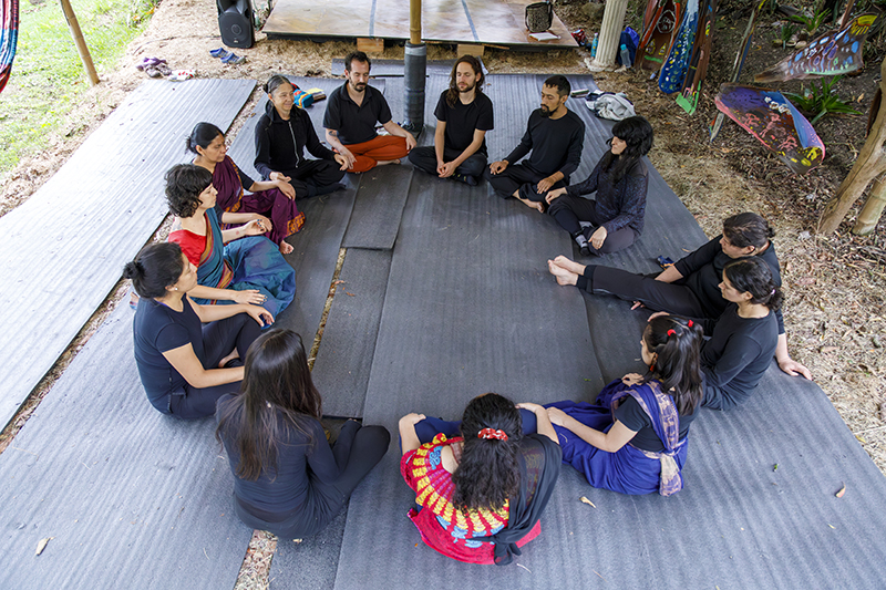 A group of people sit in a circle on large mats beneath an open-air shelter in a forested setting, participating in a group discussion or reflective gathering surrounded by colorful fabrics and natural greenery.