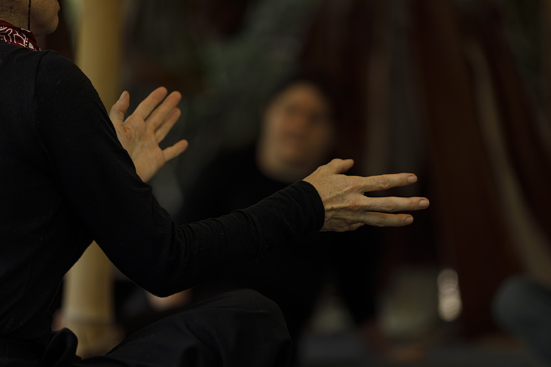 A close-up image focuses on a person's outstretched hands mid-gesture during a discussion or expressive activity, with another seated participant softly blurred in the dimly lit background.