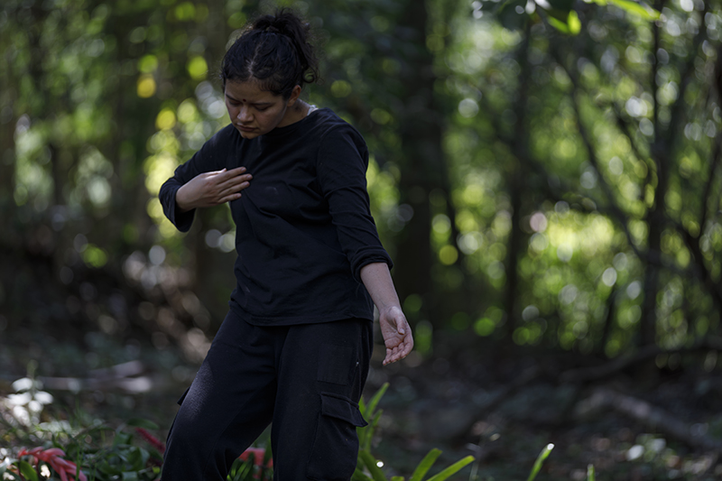 A person dressed in black performs a deliberate movement sequence in a forest setting, with one hand resting near the chest and the other extended downward amid softly blurred greenery.