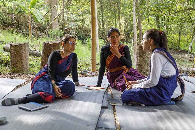 Three people seated on outdoor mats in a wooded setting engage in a focused discussion, with notebooks placed nearby and natural sunlight filtering through the trees.