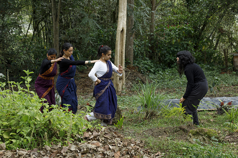 Four participants engage in a collaborative movement exercise in a wooded garden, with three people advancing in coordinated poses toward another participant who responds with a contrasting stance amid dense greenery.