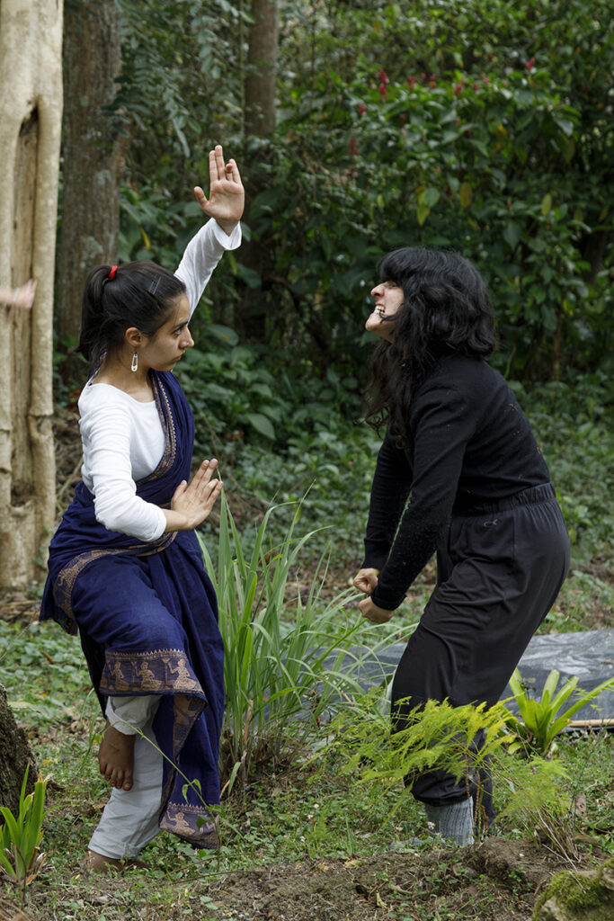 Two people engage in a dynamic movement exercise in a lush garden setting, with one person holding a stylized pose while the other responds through expressive body language amid surrounding greenery.