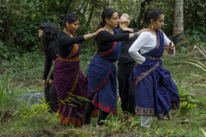 Five people in layered blue, purple, black, and white garments perform a coordinated expressive movement sequence outdoors in a lush green area surrounded by trees and foliage.