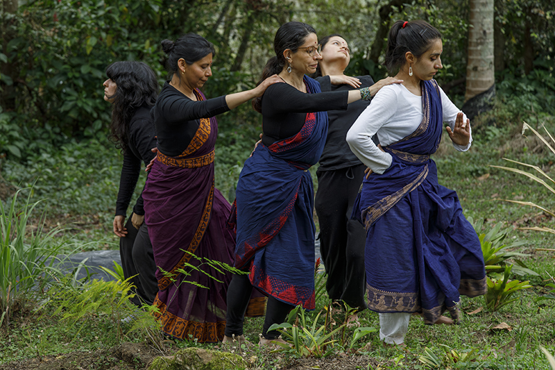 Five people in layered blue, purple, black, and white garments perform a coordinated expressive movement sequence outdoors in a lush green area surrounded by trees and foliage.