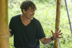 A participant in a black shirt demonstrates an expressive movement or theatrical gesture beneath a bamboo shelter, with one hand gripping a post and the other extended against a backdrop of sunlit greenery.