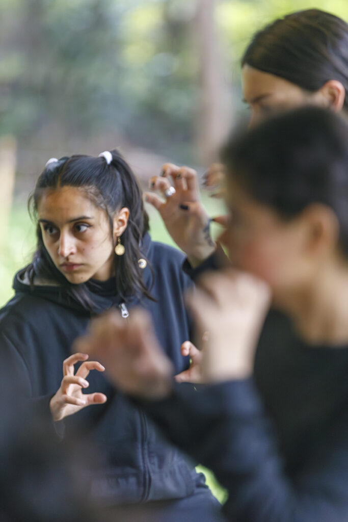 A participant in dark clothing makes a focused hand gesture while observing an outdoor group exercise, framed by other blurred participants in the foreground and background.