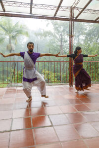 Four participants engage in a collaborative movement exercise in a wooded garden, with three people advancing in coordinated poses toward another participant who responds with a contrasting stance amid dense greenery.