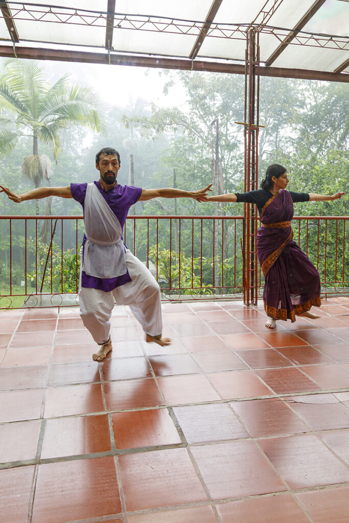 Four participants engage in a collaborative movement exercise in a wooded garden, with three people advancing in coordinated poses toward another participant who responds with a contrasting stance amid dense greenery.