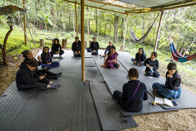 A group of participants sits in a wide circle on mats beneath an open-sided shelter in a forested environment, engaged in a collaborative discussion or learning session surrounded by trees, hammocks, and natural greenery.