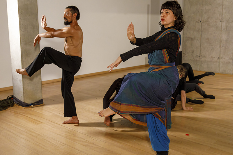 Several people perform expressive, dance-like poses in a spacious studio, with two participants balancing on one leg in the foreground while others move close to the floor in the background.