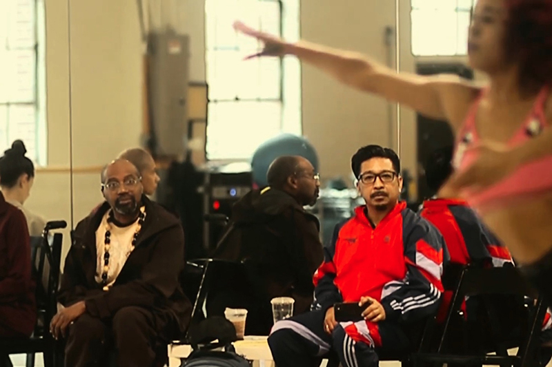 Ronald and Arcell sit and observe a dance rehearsal in a mirrored studio while a performer moves across the foreground with an outstretched arm.