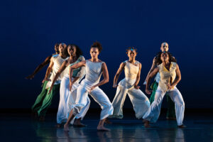 A group of dancers in flowing white and green costumes perform a synchronized contemporary dance on a stage illuminated by deep blue lighting.