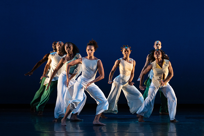 A group of dancers in flowing white and green costumes perform a synchronized contemporary dance on a stage illuminated by deep blue lighting.