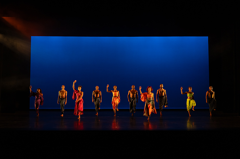 A line of dancers in colorful flowing costumes performs a synchronized movement across a dark stage, dramatically illuminated against a vivid blue backdrop.