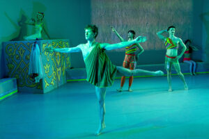 A group of performers in brightly colored costumes pose and dance across a stage bathed in blue-green light, with one dancer balancing on one leg in the foreground while others strike stylized poses around the set.