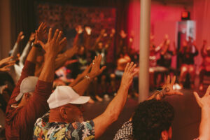 Participants seated in a circle raise their hands during an interactive community gathering in a warmly lit indoor space with red ambient lighting.