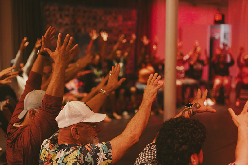 Participants seated in a circle raise their hands during an interactive community gathering in a warmly lit indoor space with red ambient lighting.