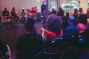 A group of people sit in a circle during an indoor community discussion or workshop, viewed from behind several attendees under soft purple and blue lighting.