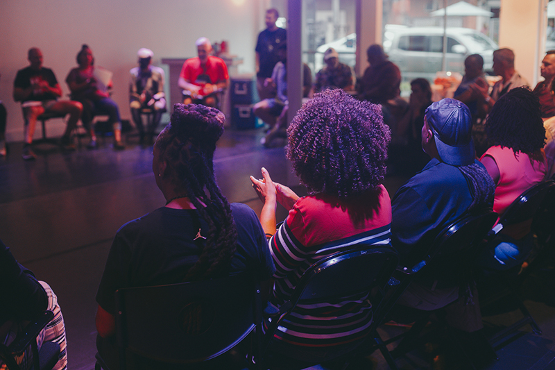 A group of people sit in a circle during an indoor community discussion or workshop, viewed from behind several attendees under soft purple and blue lighting.