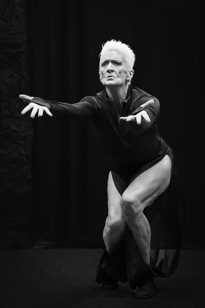 Andrea Labbee, in a dark stage setting, crouches with arms extended forward in a striking dance pose, illuminated by dramatic black-and-white lighting.
