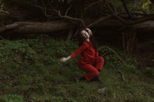 Erin is dressed in a vivid red outfit and strikes an expressive pose in a dimly lit forest clearing, framed by lush greenery and tangled fallen tree branches.