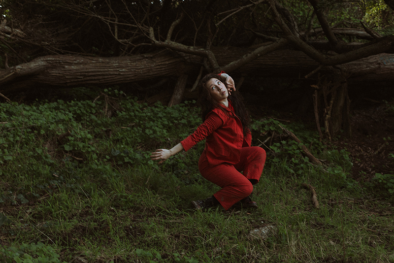 Erin is dressed in a vivid red outfit and strikes an expressive pose in a dimly lit forest clearing, framed by lush greenery and tangled fallen tree branches.