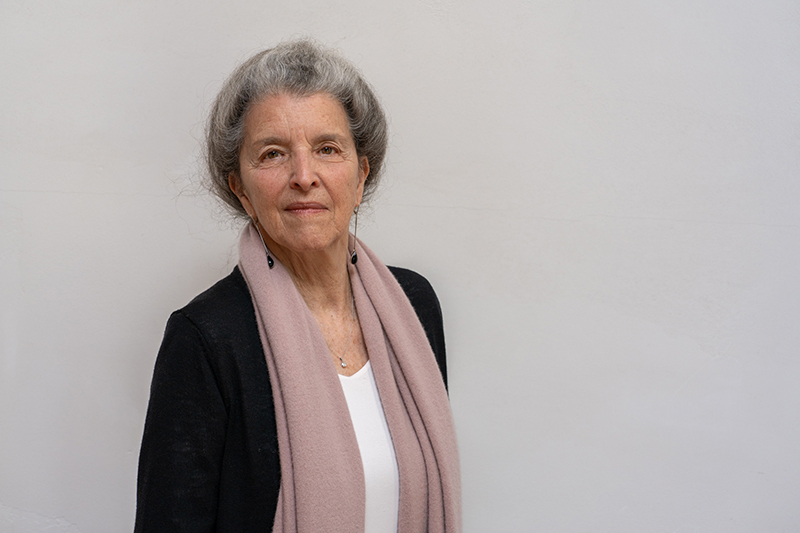 A professionally composed portrait shows a Janet with gray hair wearing a dark cardigan, light top, and soft pink scarf, standing against a plain light-colored wall.