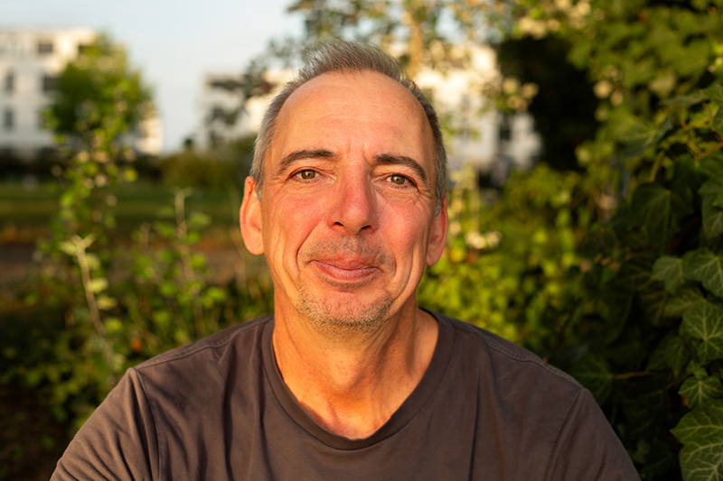 Copilot said: A close-up outdoor portrait shows Jens in a dark T-shirt looking toward the camera, illuminated by warm golden-hour sunlight with lush green foliage and softly blurred buildings in the background.