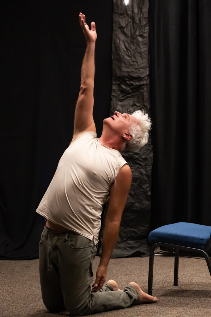 Octavio kneels on the floor beside a chair on a dimly lit stage, stretching upward with one arm extended toward the ceiling in a dramatic movement pose.